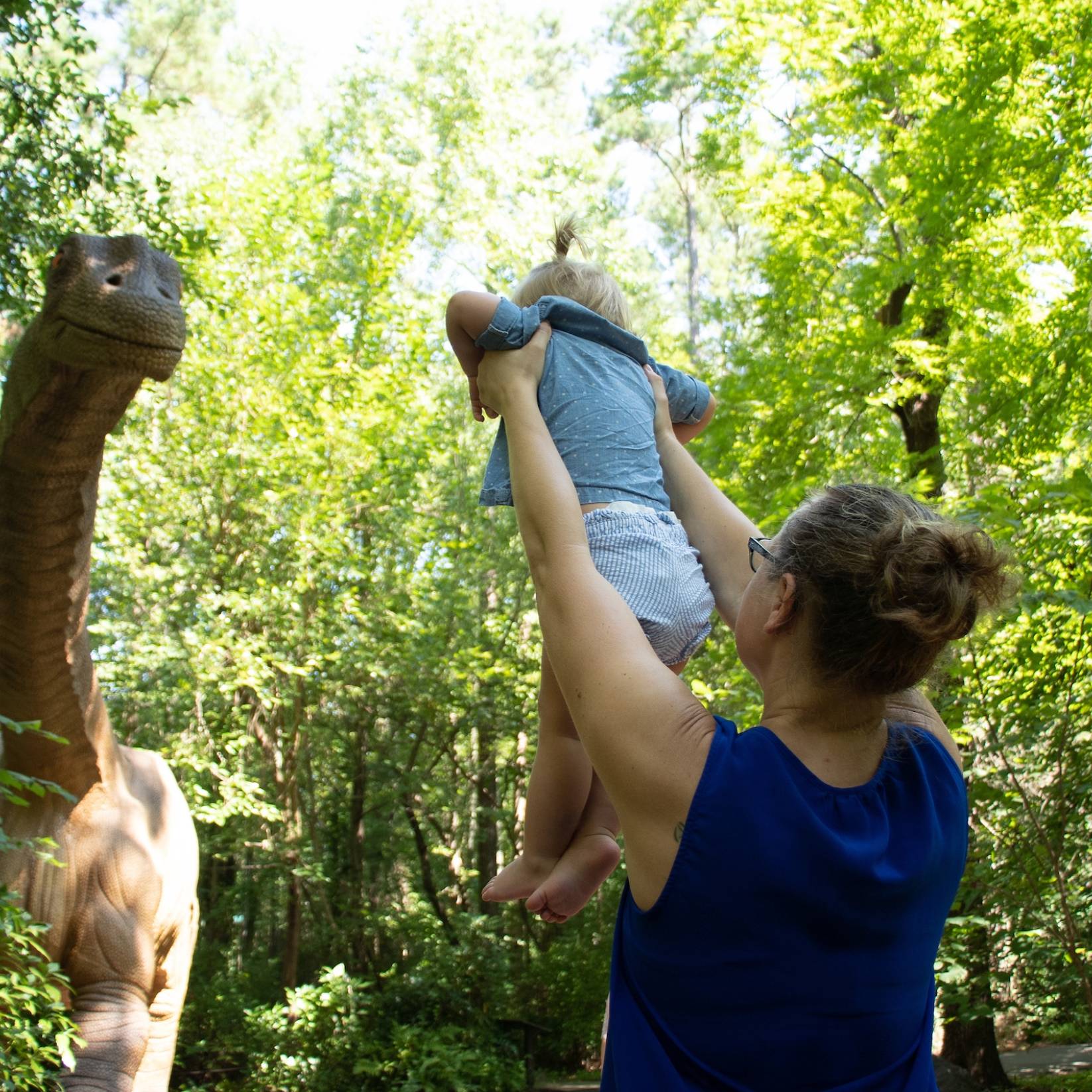 A woman holds up a baby to a life-size dinosaur replica at the Museum of Life and Science